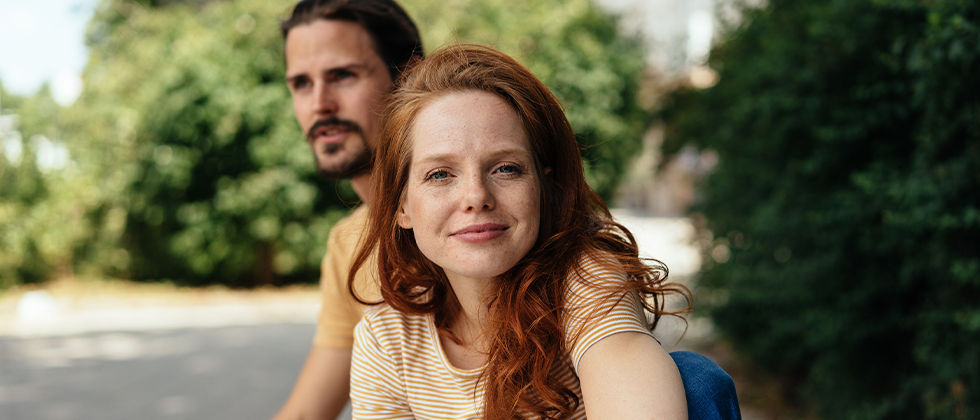 A couple sitting down, the woman in focus looking to camera, the man behind her
