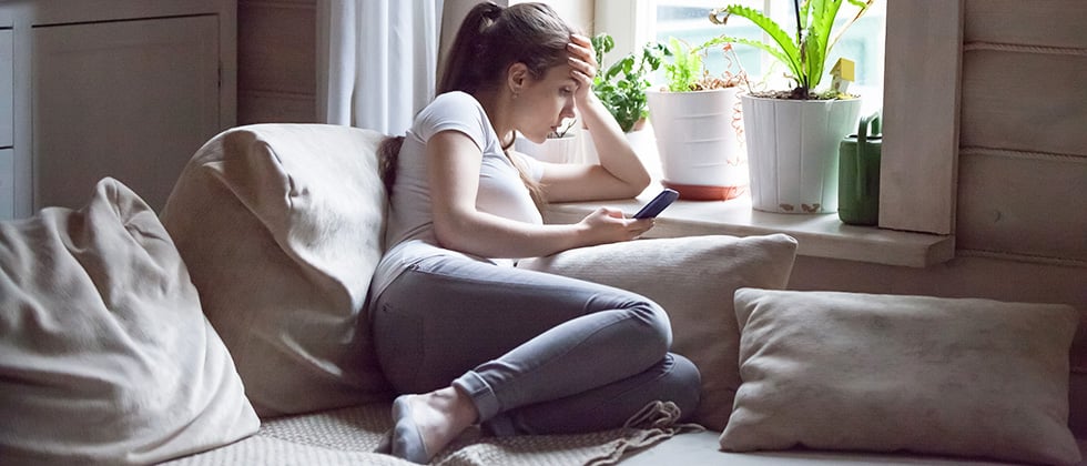 A woman sits alone on a living room sofa, looking at her phone