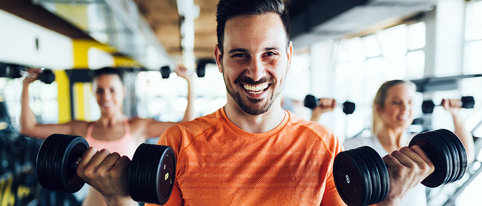 A man in a gym looks to camera while holding a dumbbell weight in each hand