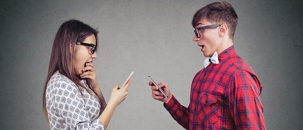 A couple face each other, looking surprised while staring at their phones
