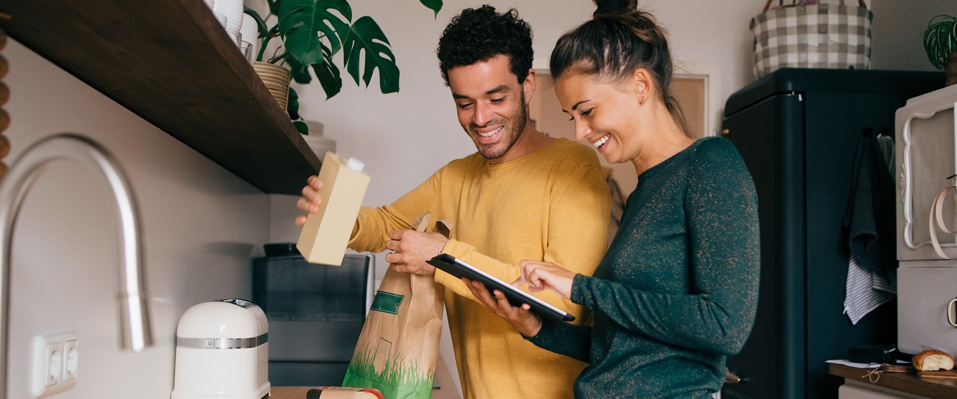 A man and a woman are standing in the kitchen unpacking gorcerie bags. The woman is searching for something on her iPad. They are laughing together and it seems like they are about to cook together.