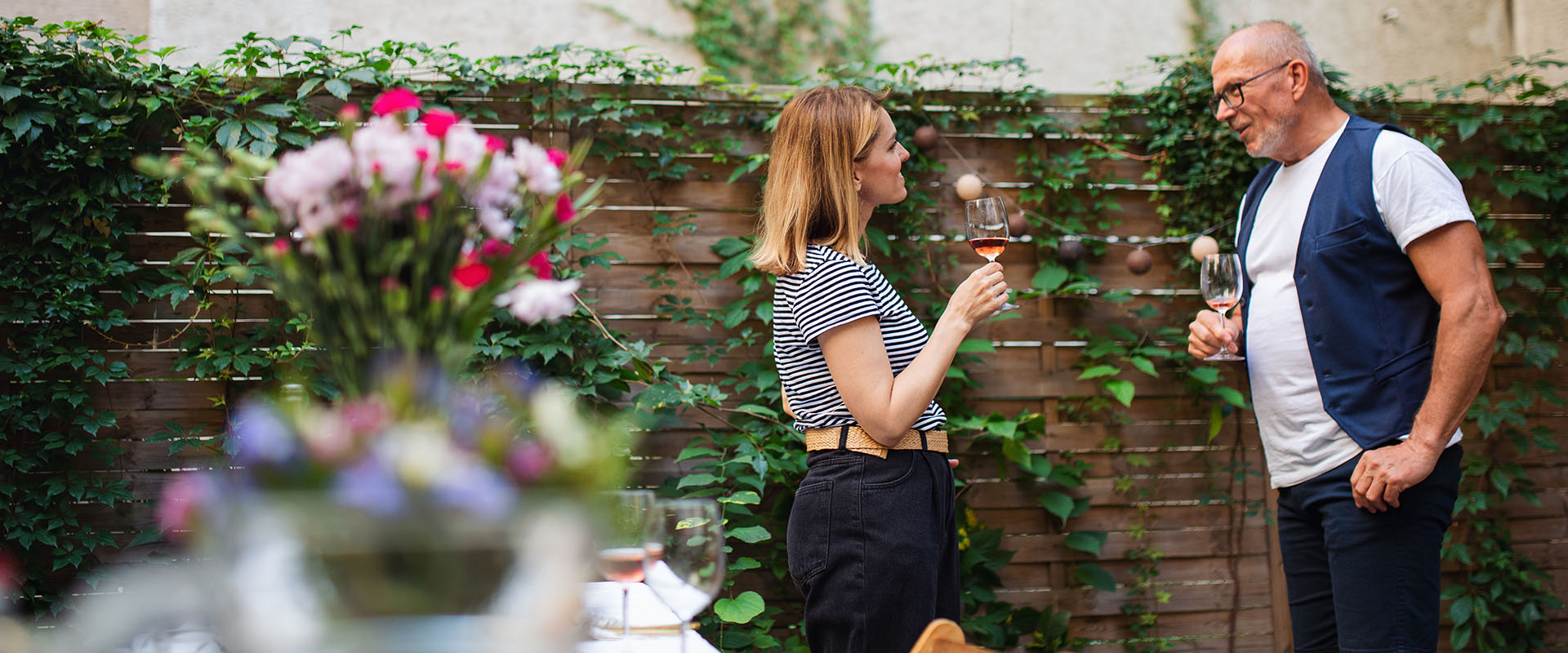 A woman and a man standing in a garden with wine glasses in their hands. They seem to have a good conversation.