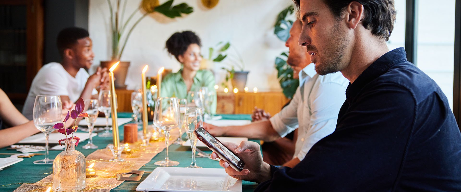 Mid-aged people sitting around a table having a dinner party together. In focus is a man distracted by his smartphone where he looks at social media with a picture of a woman.