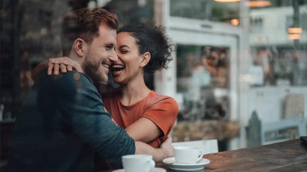 A couple in their 30s laughing and hugging in a coffeeshop