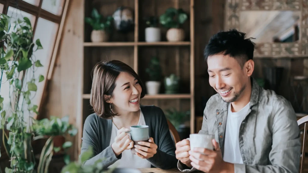 Asian couple sitting in a comfy café enjoying a cup of coffee and laughing together.