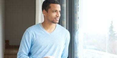 A young guy staring out a window holding coffee and smiling