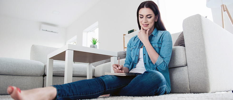 A woman sits on the living room floor in front of a sofa, writing a list