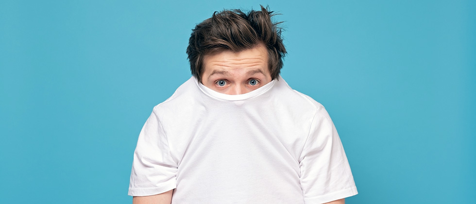 A man looks to camera having pulled the collar of his t-shirt up around his nose