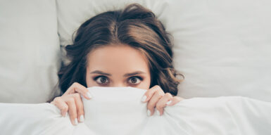 A woman in bed looking scared with the covers partly over her face