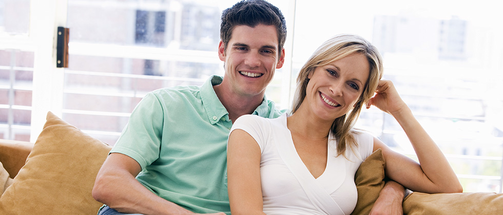 A young man and slightly older woman look to camera from their sofa