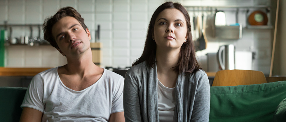Bored-looking couple sit together on a sofa, staring forward vacantly