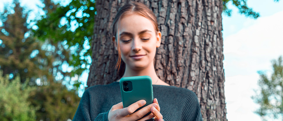 A young woman stands in front of a tree, sending a message on her phone