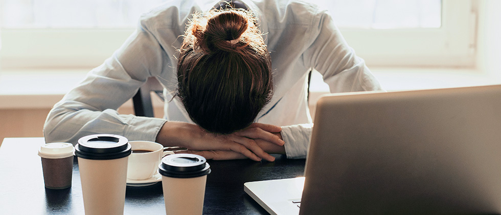 A woman slumps in front of a computer and several coffee cups, head resting on her hands