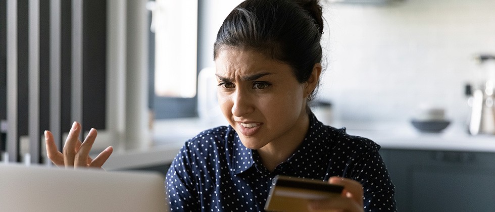 A woman stares at her laptop screen in anger, holding a credit card in one hand