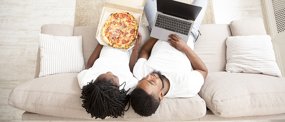 A couple sit together on a sofa, viewed from above. The woman has a pizza on her lap, the man a laptop computer