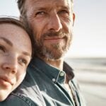 A couple is on the beach and the woman is leaning her head on the mans' shoulder. The woman seems to be younger than the man.