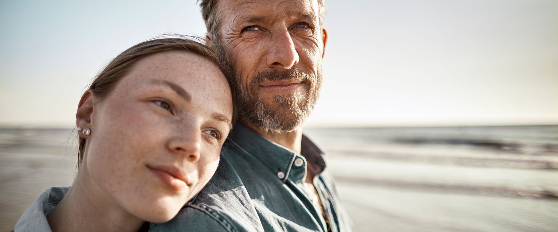 A couple is on the beach and the woman is leaning her head on the mans' shoulder. The woman seems to be younger than the man.