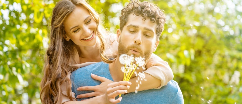 A woman on her date's back holding dandelions while he blows on them