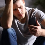 Worried and upset man sitting on the ground looking at his phone