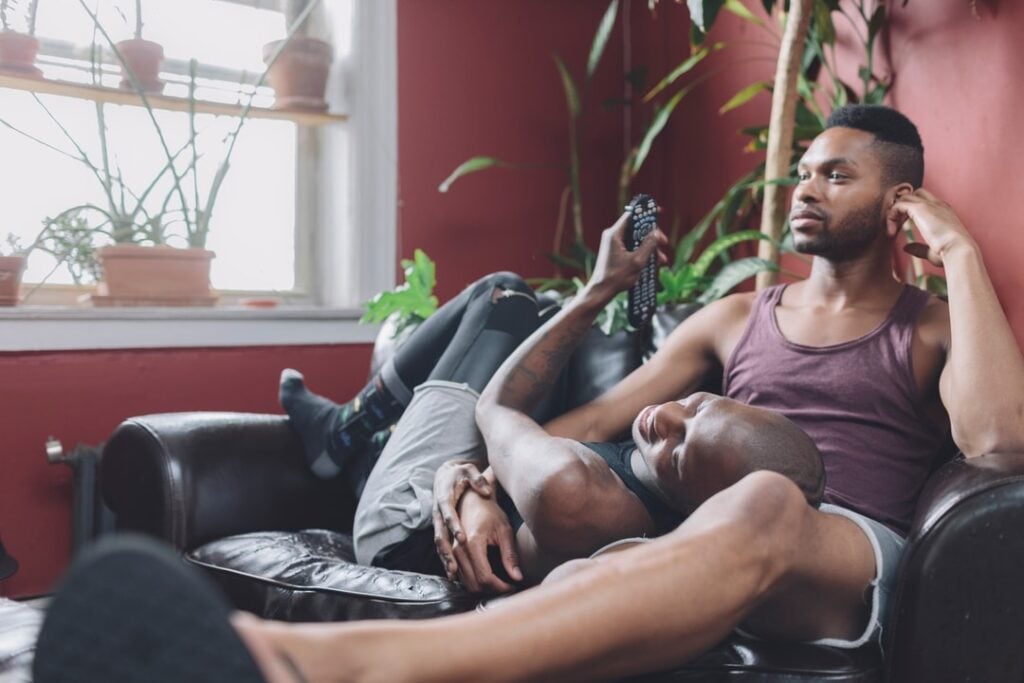 A couple laying on the couch together, one man holds the remote control and the other is watching television.