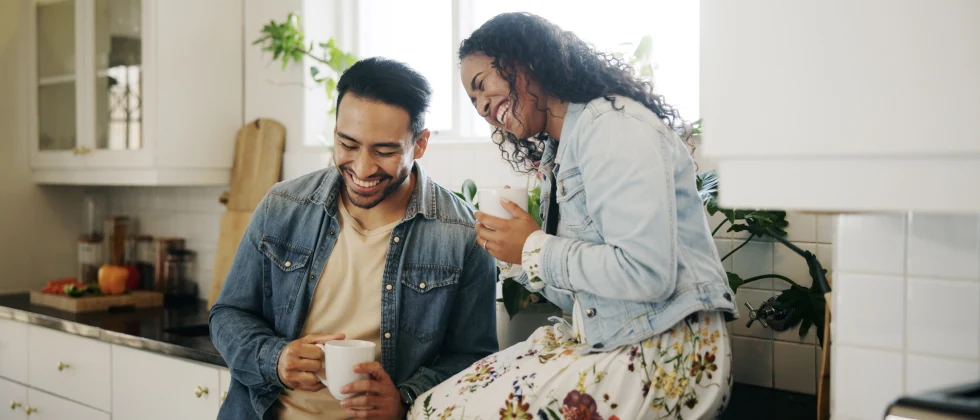 A couple sitting on the kitchen corner holding cups and laughing.
